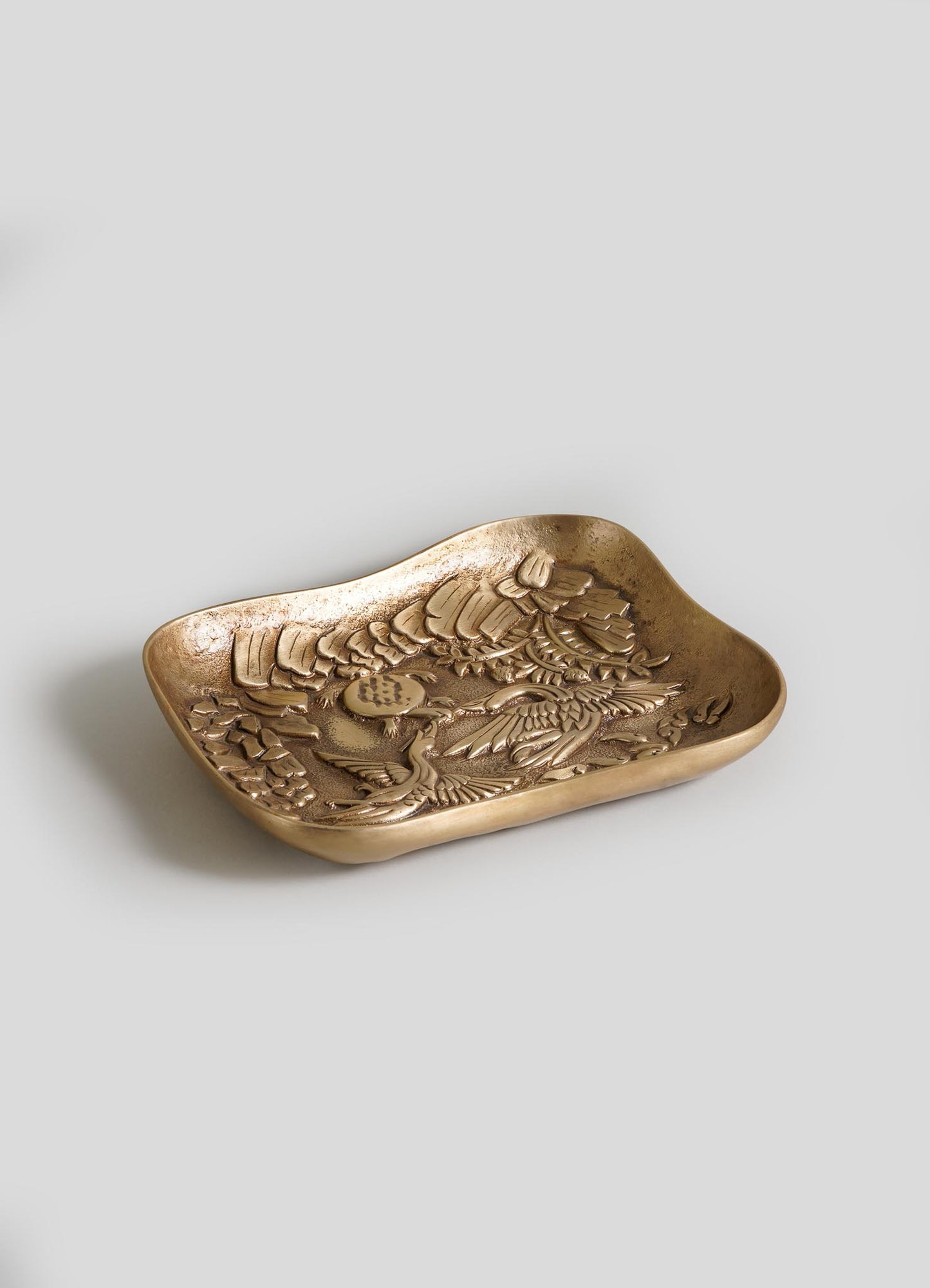 The VIYA Tortoise & Crane Trinket Bowl is a small rectangular brass catch-all tray with curved edges and an embossed bird and floral design, inspired by jungle fable decor, shown against a plain light grey background.