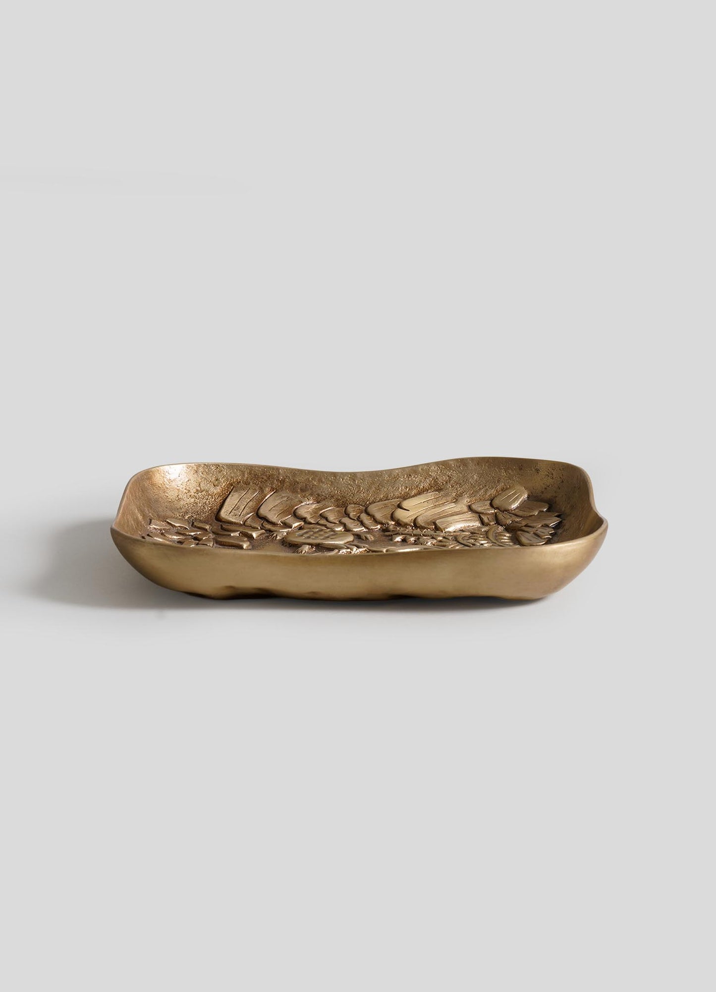 The VIYA Tortoise & Crane Trinket Bowl is a rectangular, shallow brass tray with wavy edges and a raised leaf pattern, shown against a plain light gray background.