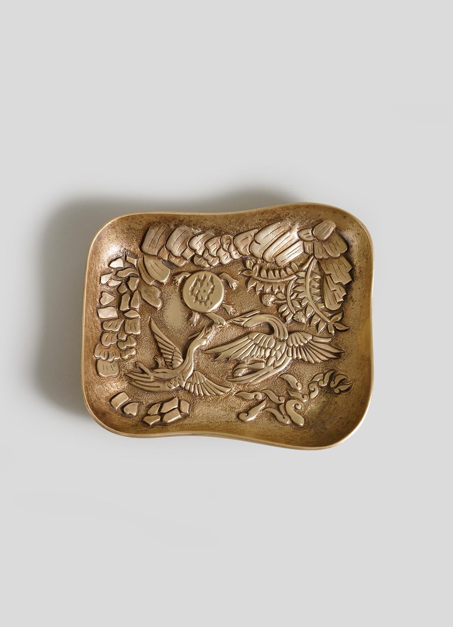 The VIYA Tortoise & Crane Trinket Bowl is a square bronze tray with curved edges, embossed with a crane among flowers—an elegant touch of jungle-inspired decor—shown on a plain gray background.