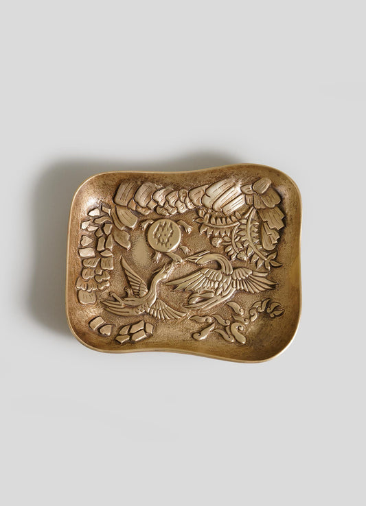 The VIYA Tortoise & Crane Trinket Bowl is a square bronze tray with curved edges, embossed with a crane among flowers—an elegant touch of jungle-inspired decor—shown on a plain gray background.