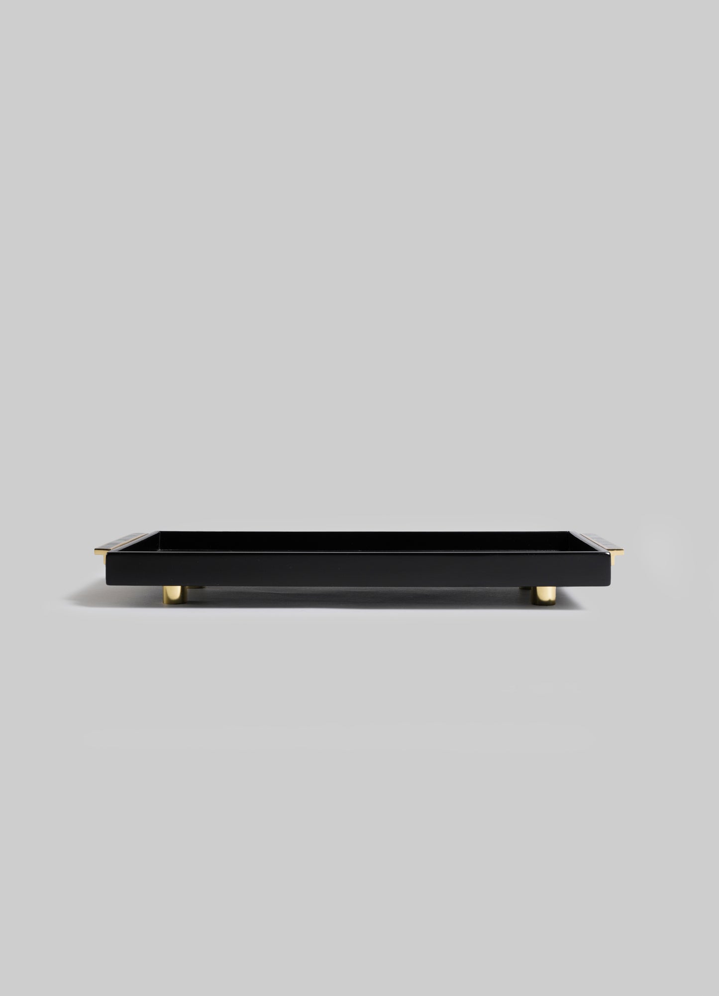 The Plaza Vanity Tray by VIYA is a rectangular black tray with gold feet and corner accents, channeling Art Deco glamour. Styled on a light gray background.