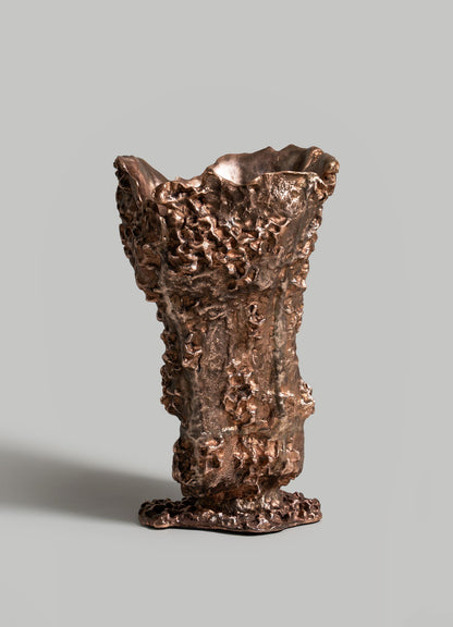 The VIYA Red Earth Vase features a bronze, textured surface with irregular edges and a rugged, organic form. It stands upright on a smooth gray background, reflecting forest-inspired artistry.
