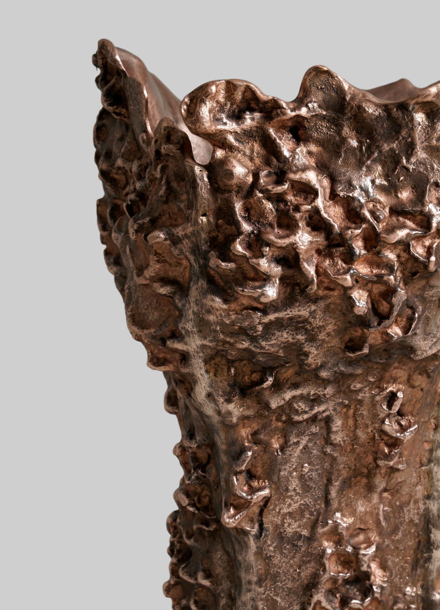 A close-up of the VIYA Red Earth Vase with a rough, organic, textured surface and irregular, jagged edges set against a plain light gray background. The vase has a sculptural form and metallic reflective finish.