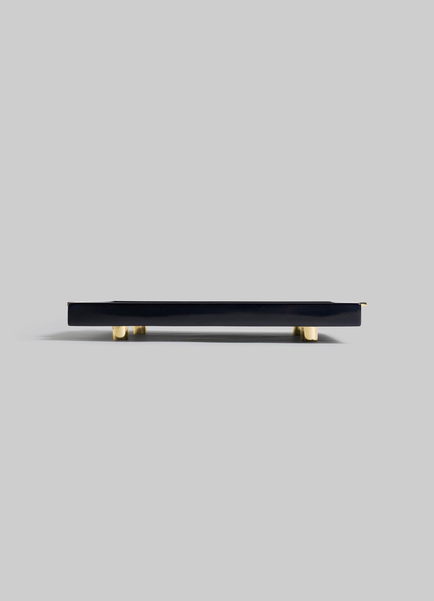 The VIYA Rivoli Black Towel Tray, featuring a sleek rectangular design with gold-colored feet, channels Art Deco elegance inspired by vintage Bombay cinema halls; showcased against a plain light gray background from a low side angle.