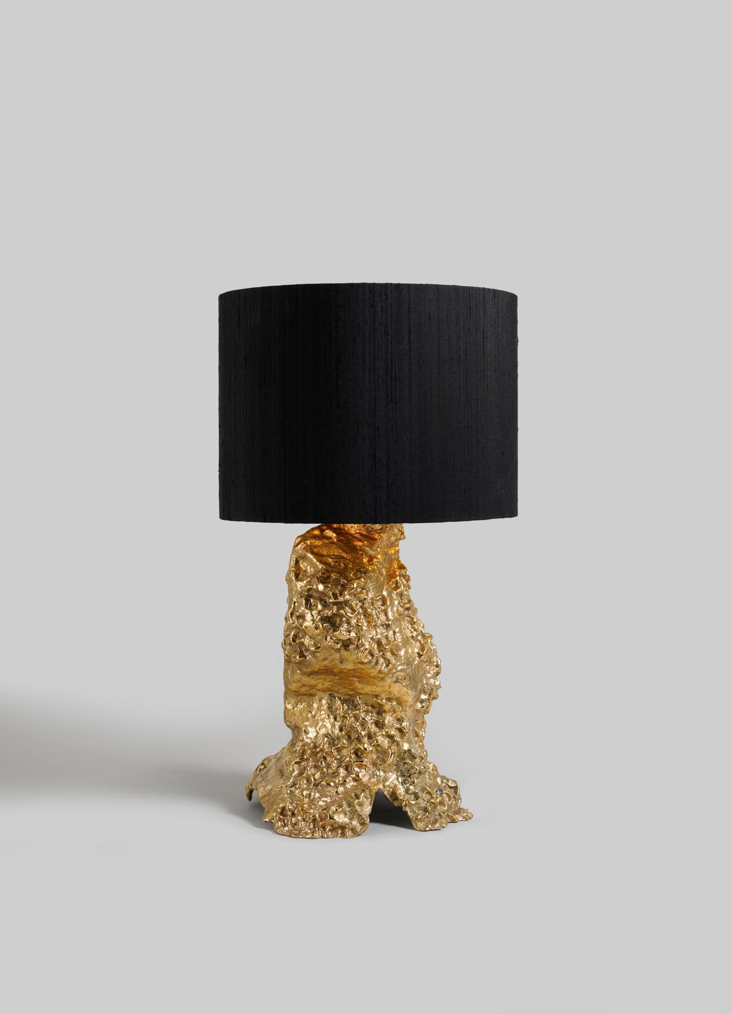 The VIYA Red Earth Table Lamp features a sculptural gold metallic base with a textured, molten appearance and is topped with a large cylindrical black shade, set against a plain grey background.