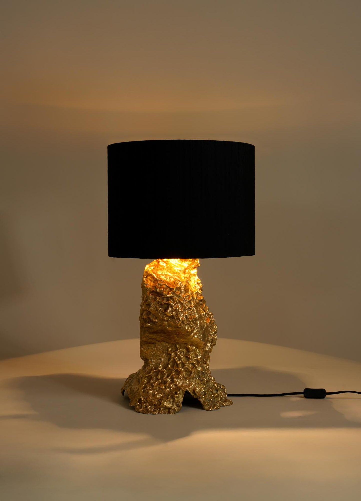 The VIYA Red Earth Table Lamp features a textured gold-toned base with a molten metal look and a large black cylindrical shade, casting warm light in neutral spaces beside sculptural pieces like the Red Earth table.