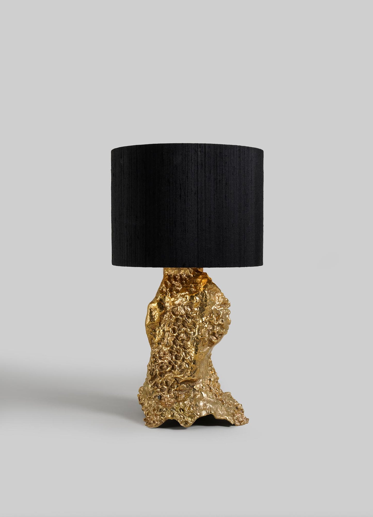 The VIYA Red Earth Table Lamp features a textured gold sculptural base and a large, round black lampshade, displayed against a plain light gray background.
