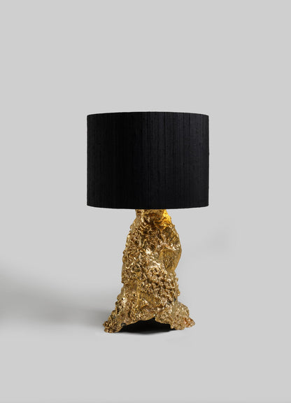 The VIYA Red Earth Table Lamp features a sculptural, textured gold base and a large cylindrical black shade, displayed against a plain light gray background.