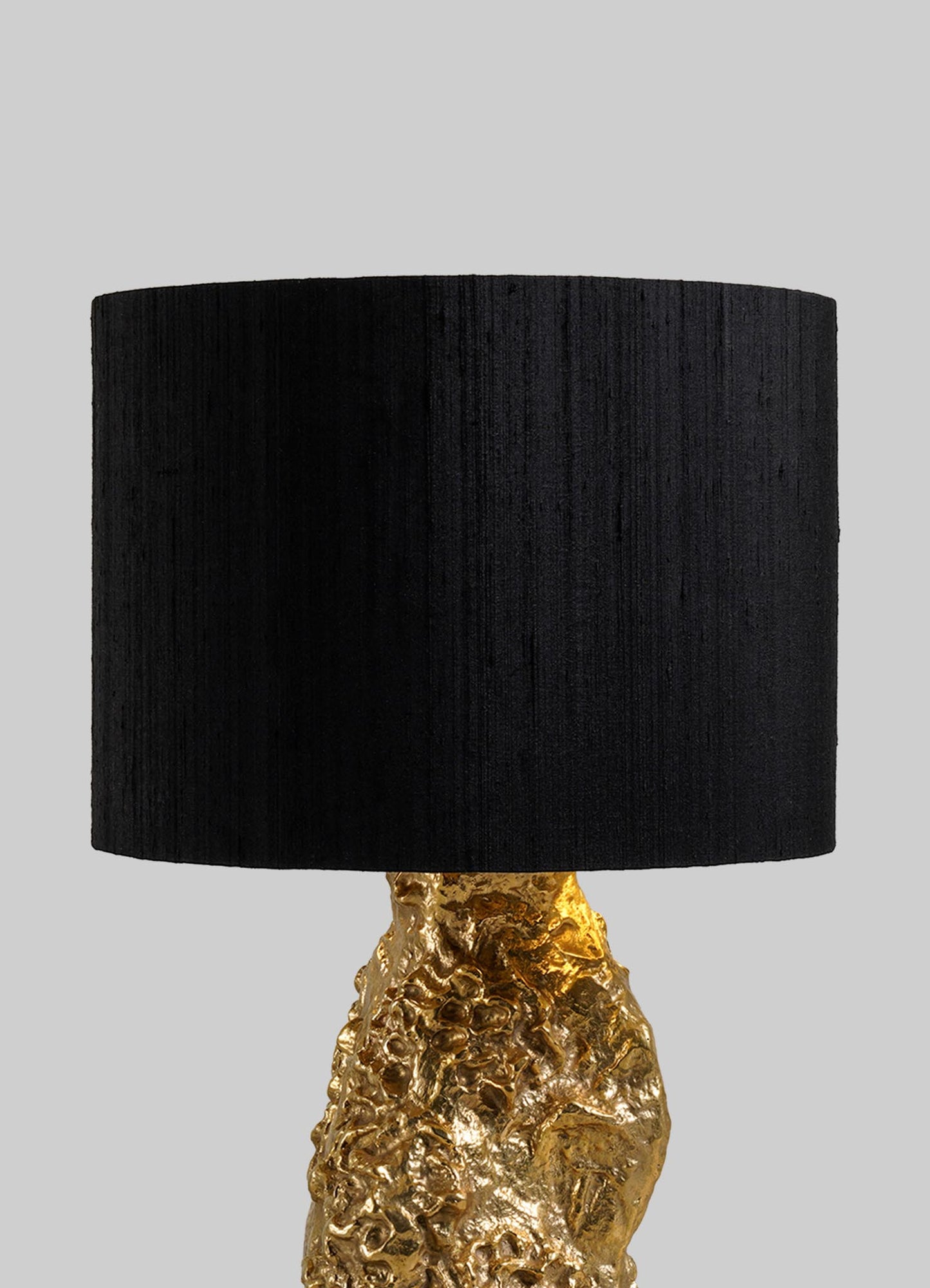 The VIYA Red Earth Table Lamp, featuring a black cylindrical shade and a textured, gold-toned base with sculptural flair, is displayed against a plain gray background.