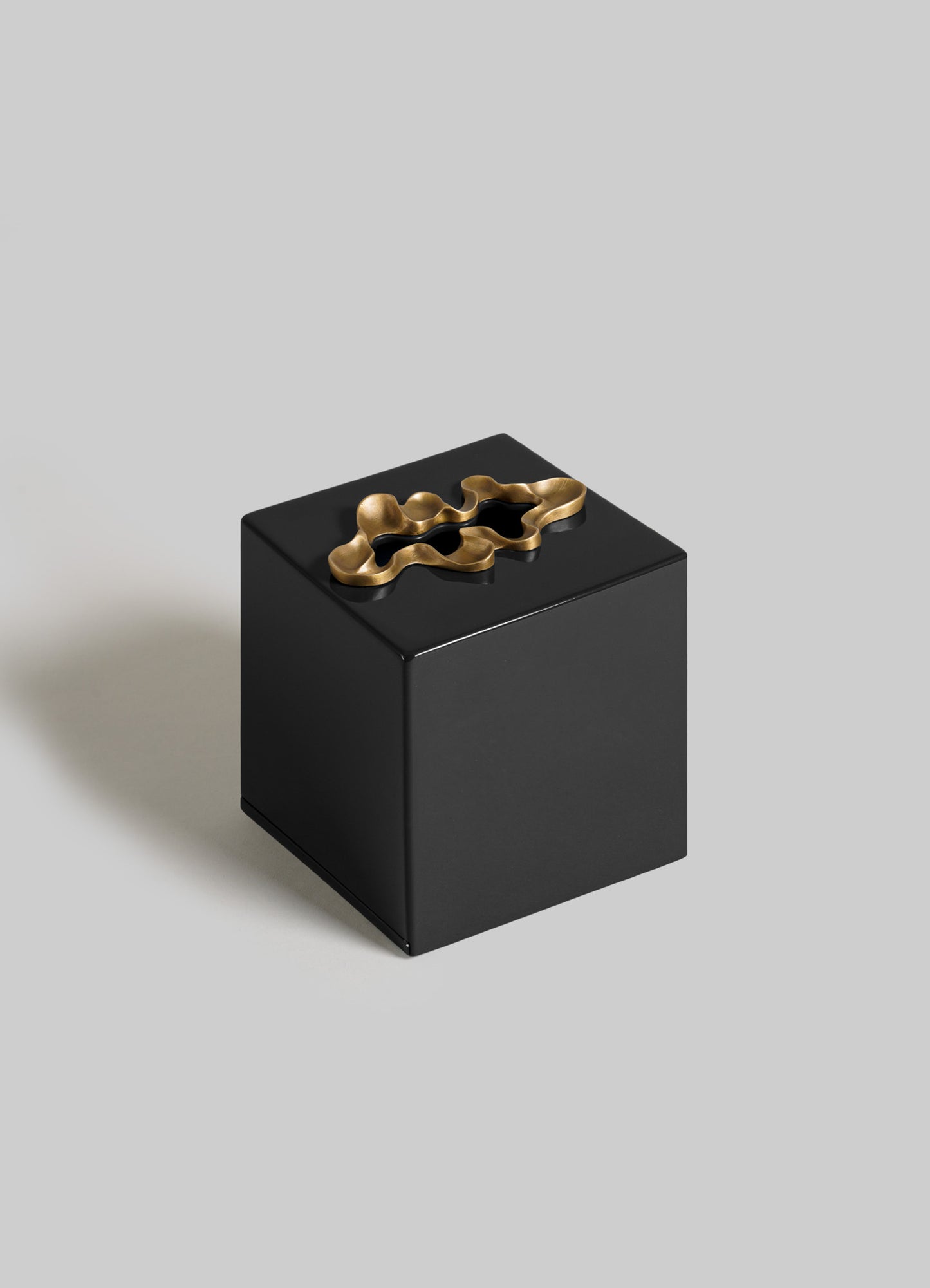 The VIYA Saffron Black Tissue Box Small features a glossy black finish and a sculptural gold metallic lid accent, echoing contemporary bronzed art, shown against a plain light gray background.