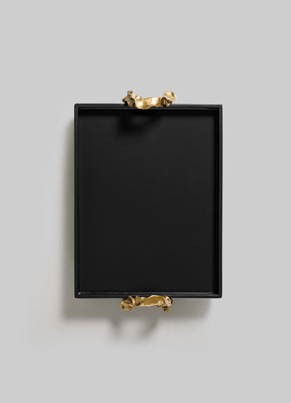The Saffron Black Towel Tray by VIYA is a square black tray featuring two sculptural gold handles on opposite sides, styled like contemporary bronzed objects and displayed on a plain light gray background.