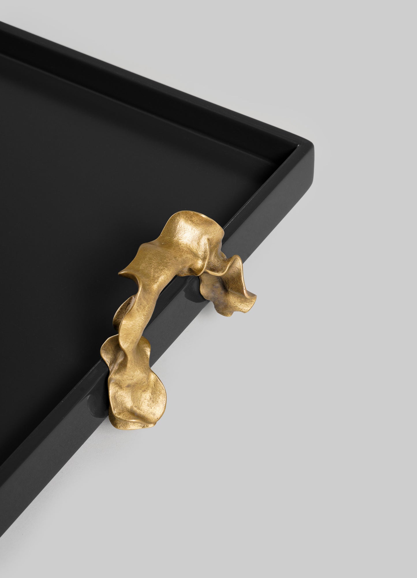 The VIYA Saffron Black Towel Tray features a black rectangular base with a sculptural gold handle shaped like a flowing ribbon, attached at the corner, and is displayed against a plain light gray background.