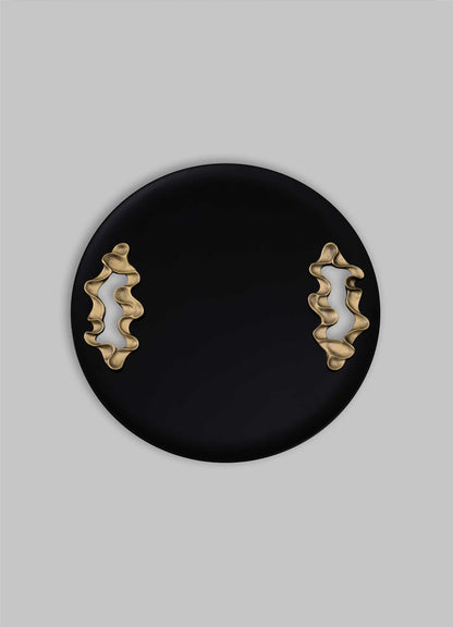 The Saffron Black Tray Large by VIYA features a round black design with two wavy gold accents on opposite sides, reminiscent of contemporary bronzed objects, displayed against a plain gray background.