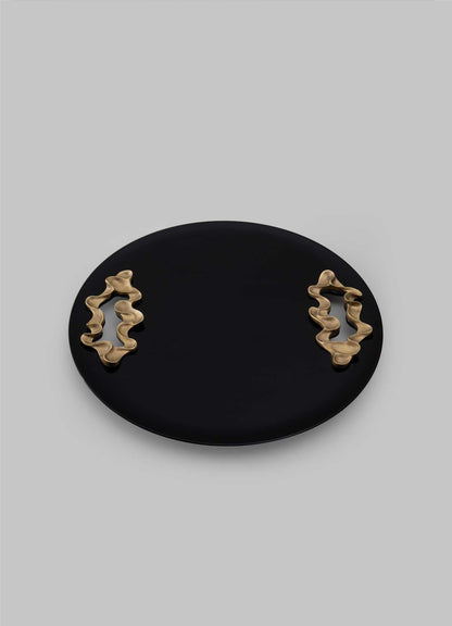 The VIYA Saffron Black Tray Large features a round, flat black design with two abstract wavy gold handles on opposite sides, evoking contemporary elegance against a plain light gray background.