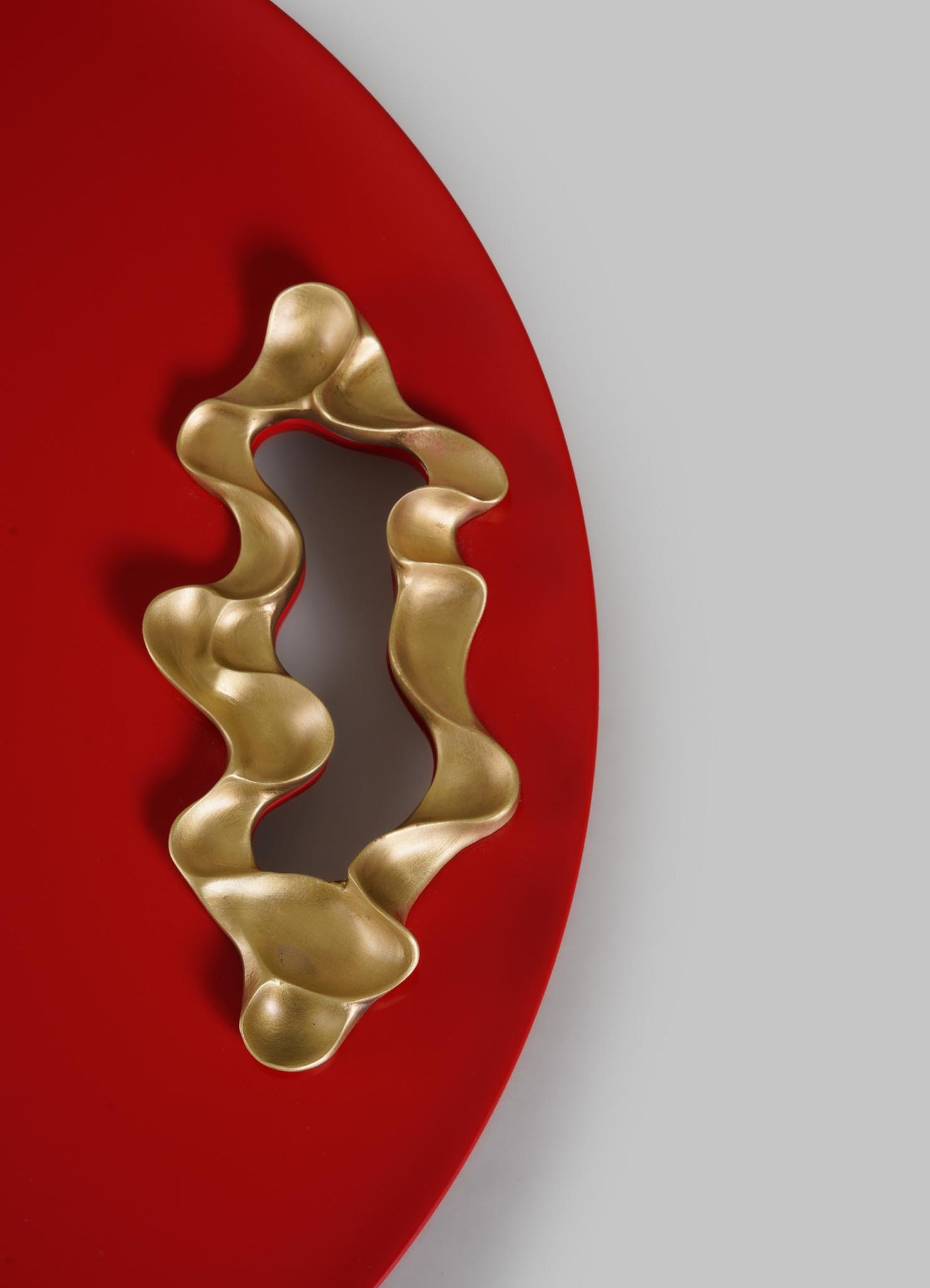 A wavy, abstract gold sculpture rests on VIYA's Saffron Red Tray Small, its round, vibrant surface recalling saffron threads. The smooth, freeform sculpture stands out against the plain light gray background.