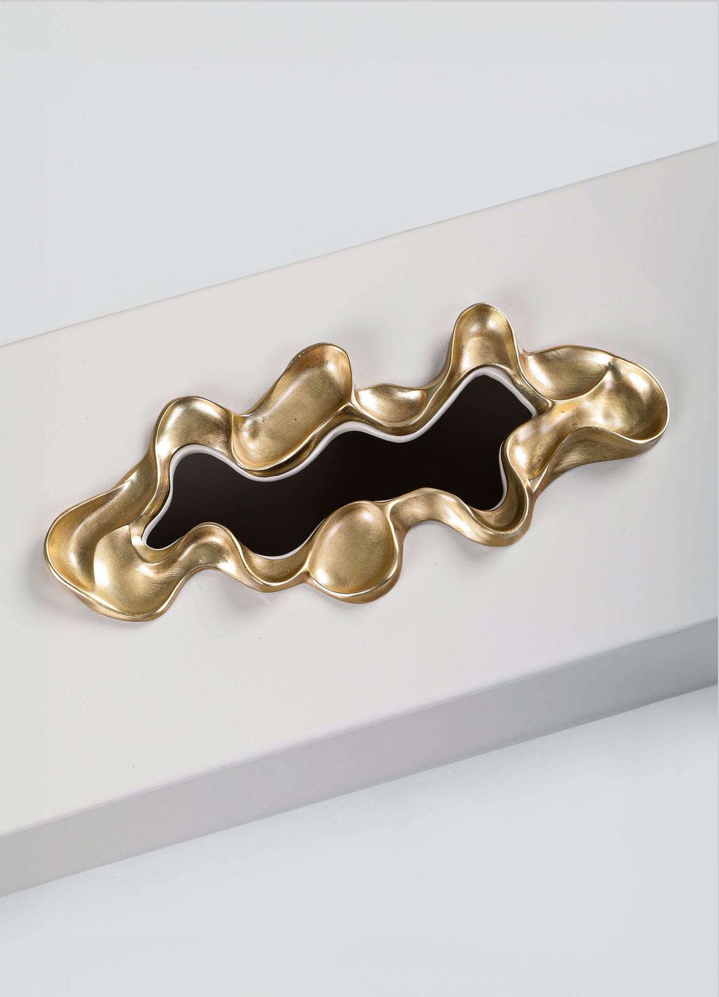 The VIYA Saffron Tissue Box Large features a sculptural gold-toned design with wavy edges and a glossy black center, evoking contemporary elegance when displayed on a sleek white rectangular surface.