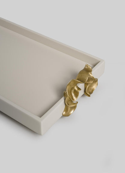 The VIYA Saffron Ecru Vanity Tray is a rectangular, off-white tray featuring a sculpted gold accent that resembles flowing leaves or draped fabric on one corner, set against a plain light gray background.