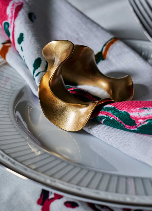 A white napkin with colorful floral patterns is held by a gold, abstract-shaped VIYA Saffron Napkin Ring, set on a white plate with ridged edges.