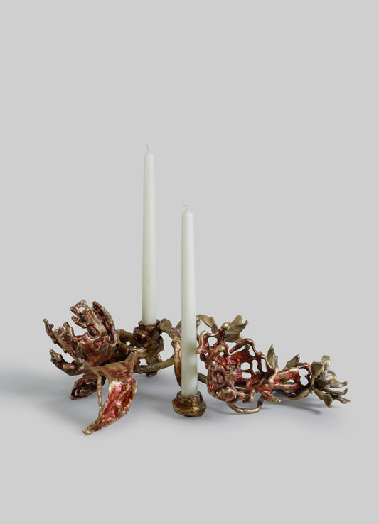 A decorative candle holder made of metal with an organic, sculptural design resembling coral or natural branches, finished in gold and reddish tones. It features two white taper candles and has a rustic, handcrafted style.