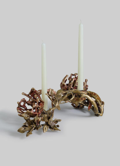 A decorative candle holder crafted from metal with an organic, floral-inspired design in bronze and reddish tones, holding two tall white candles. The piece has a handcrafted style with intricate, flowing motifs resembling leaves and flowers.