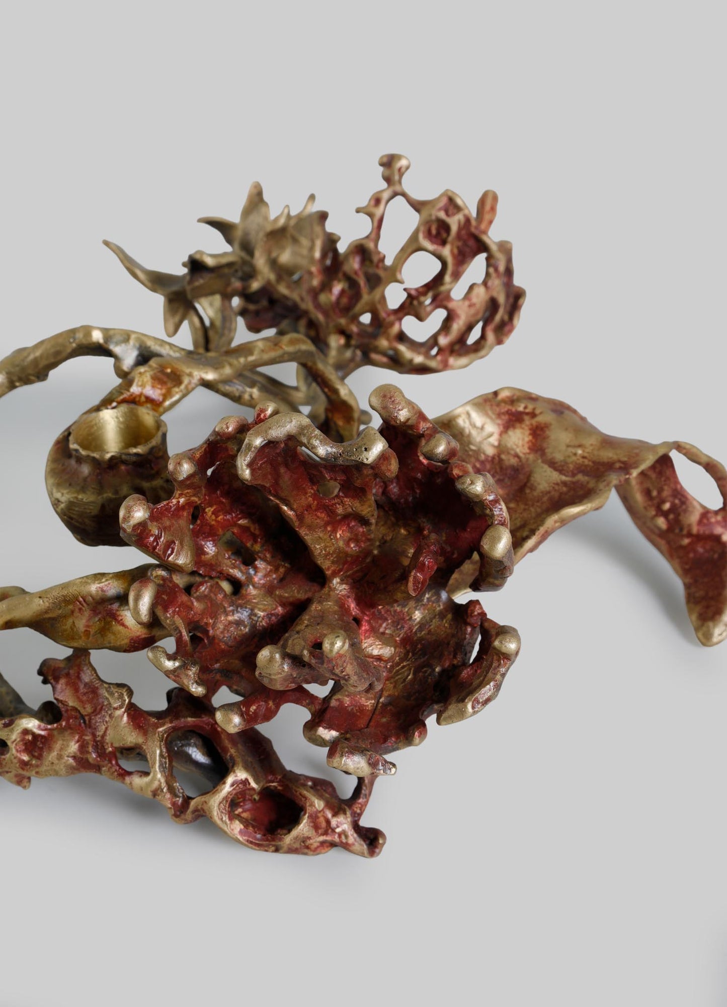 A metallic sculpture with a floral or organic motif, featuring intricate, twisting branches and blossom-like structures in a gold and reddish hue, set against a neutral background. The piece has a handcrafted, contemporary style with detailed textures and a polished finish.