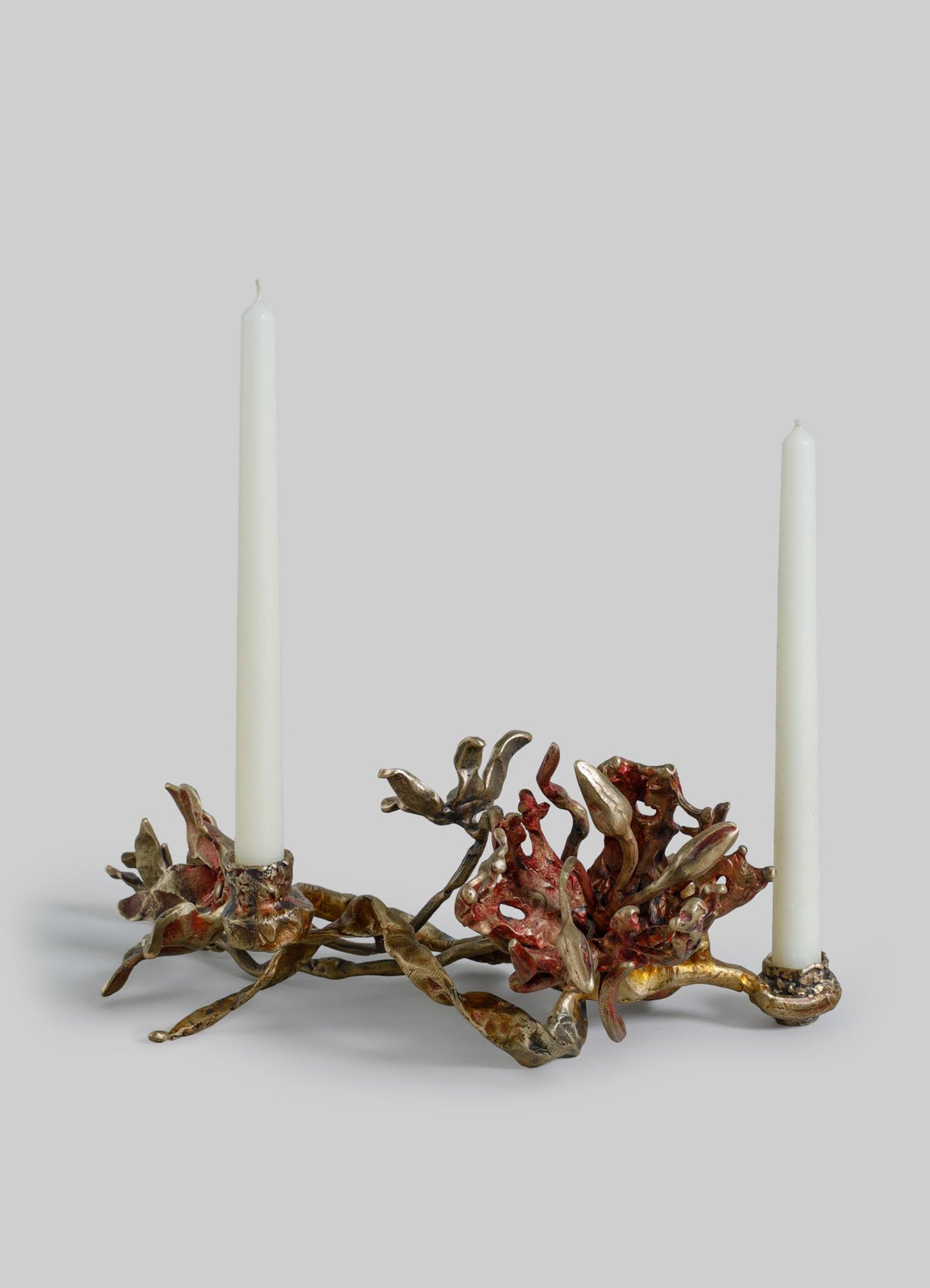 A decorative candle holder with a metallic finish resembling coral or abstract organic shapes, featuring two tall white taper candles. The design combines natural motifs with a contemporary style.