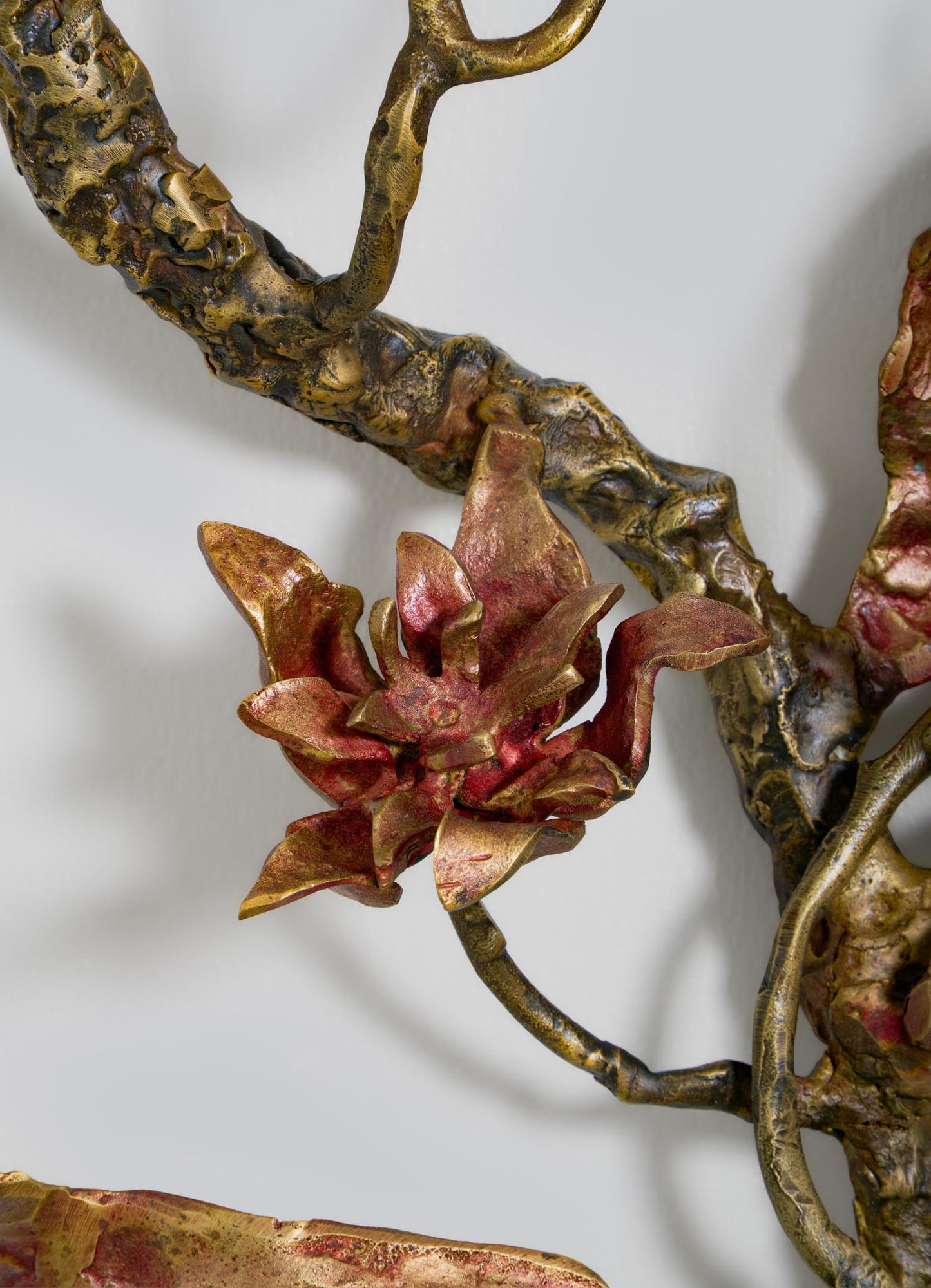 A close-up of VIYA’s Spring In Kashmir Wall Sconce B—a metallic brass and bronze sculpture with detailed red and gold flower petals on a twisting branch, serving as elegant home decor against a light gray background.