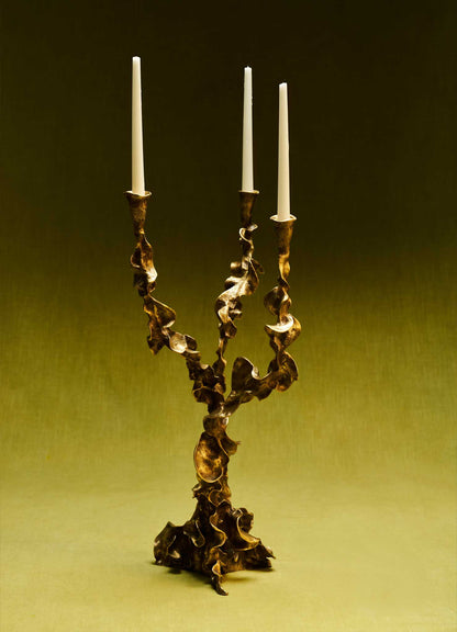 Brass candle holder with an organic, twisted design and a metallic gold finish, holding three tall white taper candles against a dark background.