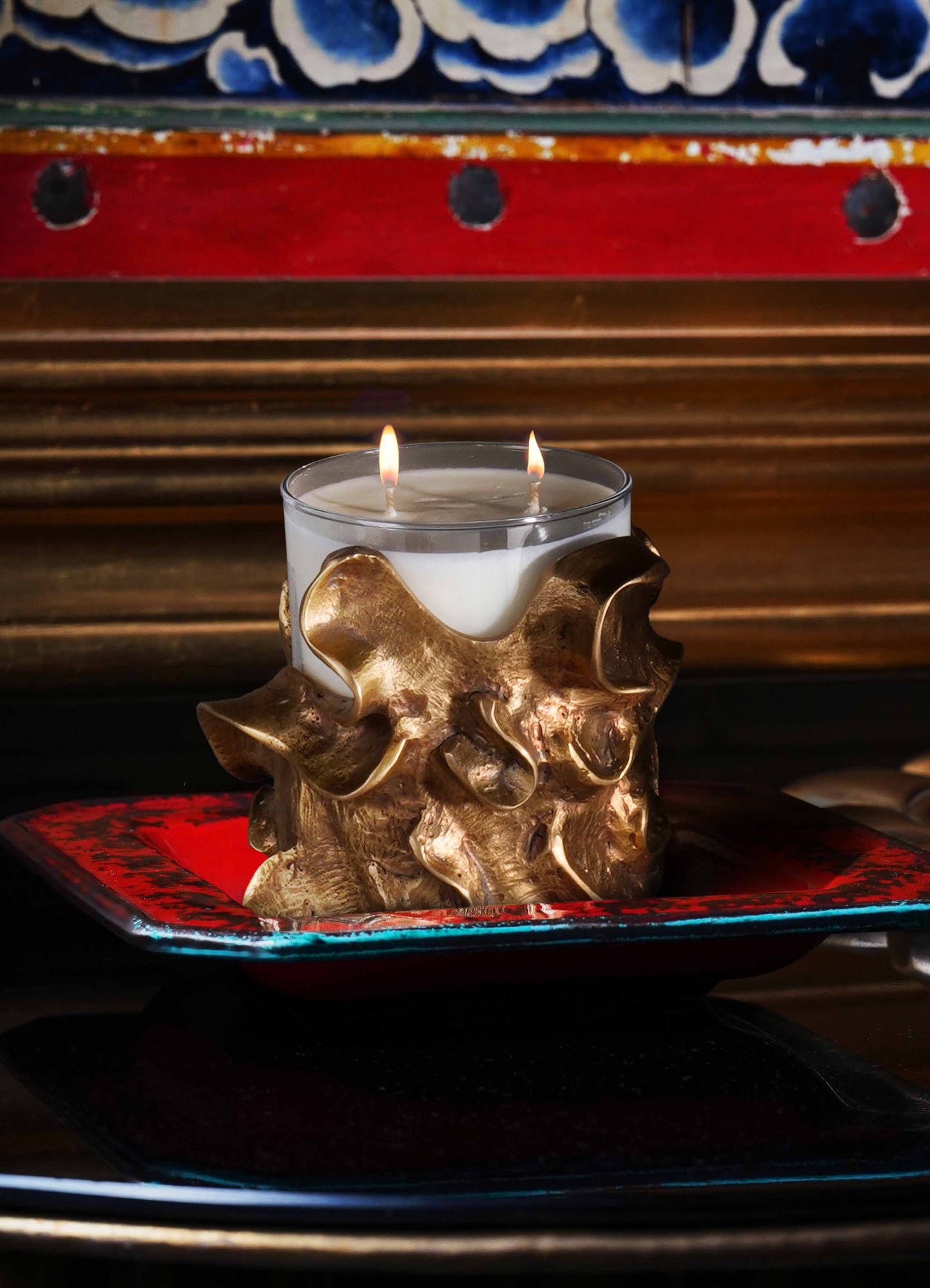A decorative candle holder with a textured metallic gold finish in irregular, organic shapes, holding a glass container with two lit candles. The candle holder is placed on a red and black glazed ceramic tray with a glossy surface.