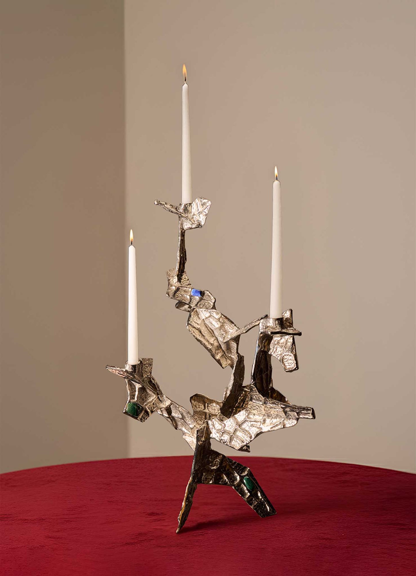 The VIYA Tutti Frutti Nickel Candelabra, featuring an abstract angular design with embedded semi-precious stones and three white candles, stands on a red velvet surface against a plain beige background.