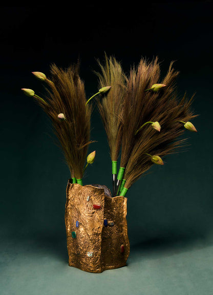 The Tutti Frutti Vessel by VIYA is a decorative gold vase featuring vibrant jewels and gemstones, filled with tall brown grass and green lotus buds, set against a dark green background.