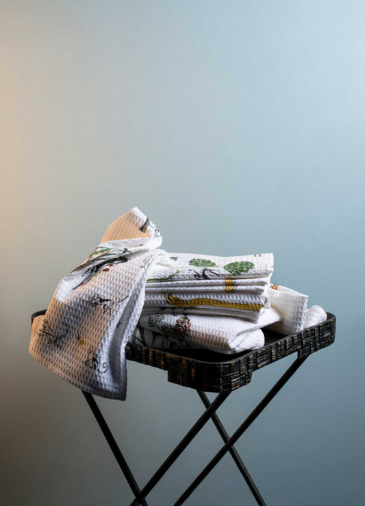 The Majestic Blackbuck Hand Towels - Set of 2 by Viya, featuring Indian wildlife-inspired floral patterns, are neatly stacked on a black metal tray against a plain light blue background, with one towel draped over the tray’s edge.