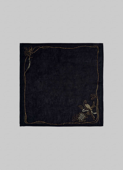 The Viya Tree Full of Parrots Hand Embroidered Black & Gold Napkin features elegant gold embroidery—a flower in the top left and a bird with branches in the bottom right—symbolizing blessings on a dark, rectangular linen.