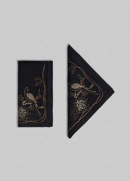 Two Viya "Tree Full of Parrots" hand-embroidered black napkins, one folded rectangularly and the other diagonally, display gold bird and floral motifs on a gray background, symbolizing blessings and good fortune.