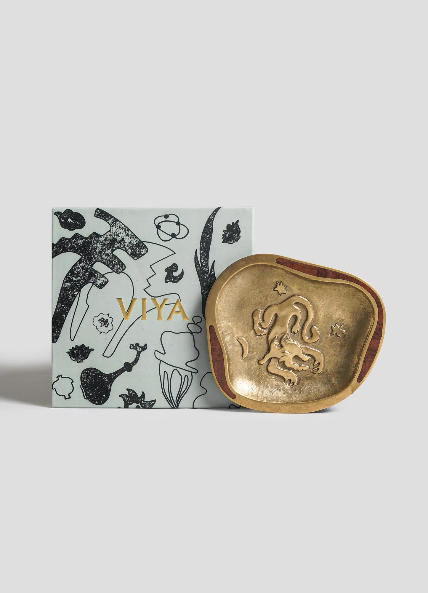 The Aries Trinket Tray by VIYA, a decorative brass dish with an abstract animal design inspired by celestial forces, is shown next to a light gray box with black line art and "VIYA" in gold lettering.