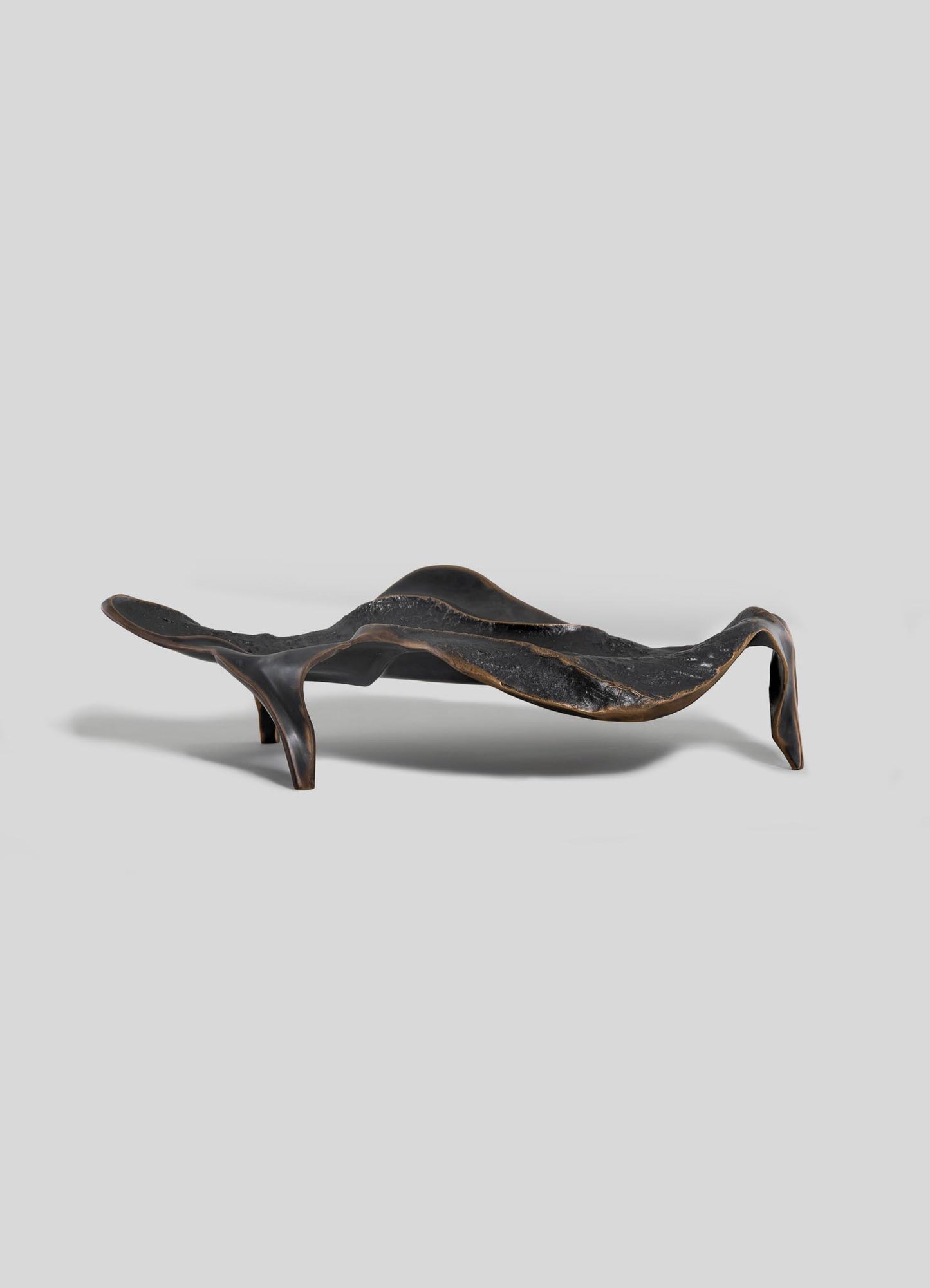 The VIYA Under The Banyan Bowl Large is a sculptural bronze piece with an organic, wavy shape and three asymmetrical legs, inspired by the banyan tree's longevity and shown against a plain light gray background.