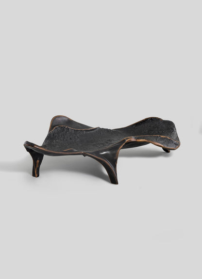 The Under The Banyan Bowl Large by VIYA is a sculptural black wooden tray with an irregular shape, three uneven legs, and textured details. Inspired by banyan trees, it features smooth, slightly glossy edges on a light gray background.
