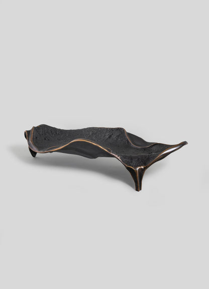 The Under The Banyan Bowl Large by VIYA is a sculptural, dark-hued tray with an organic shape and textured surface. It rests on three angled legs, featuring wavy edges with a metallic bronze finish inspired by brass symbols of longevity.