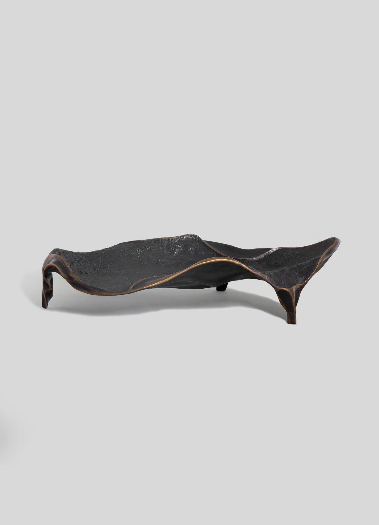 The VIYA Under The Banyan Bowl Large is a sculptural black tray with a textured surface, elevated uneven edges, and three short legs—evoking timeless brass forms—shown against a plain light gray background.