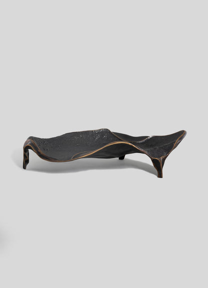 The VIYA Under The Banyan Bowl Large is a sculptural black tray with a textured surface, elevated uneven edges, and three short legs—evoking timeless brass forms—shown against a plain light gray background.