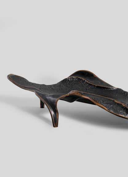 The Under The Banyan Bowl Large by VIYA features an abstract black metal form with a textured surface and wavy curves on three angled legs, evoking the enduring nature of a Banyan tree, set against a plain gray background.