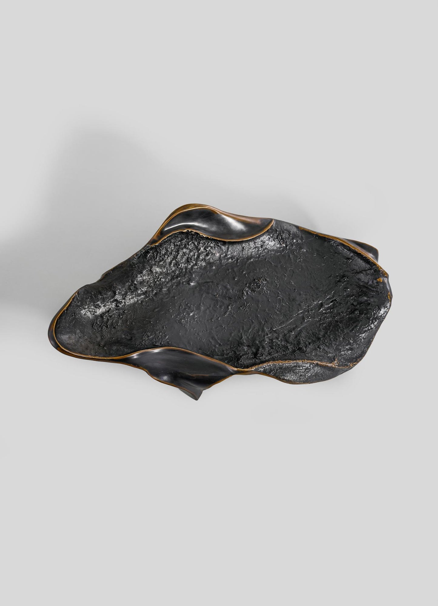 The Under The Banyan Bowl Large by VIYA features a sculptural black ceramic form with organic curves, rough textured surface, and curled edges. Subtle gold accents rim the design, displayed against a plain light gray background.