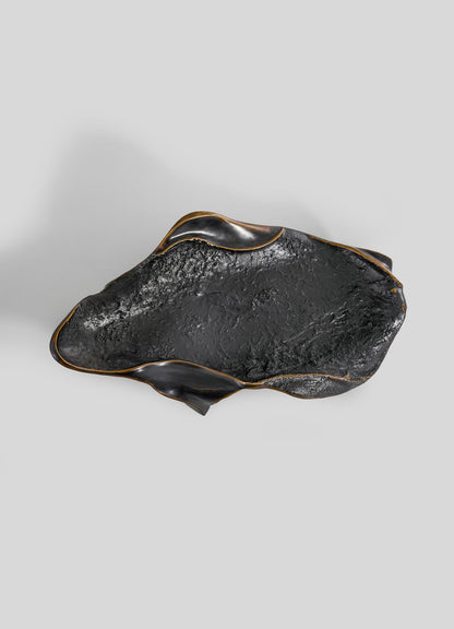 The Under The Banyan Bowl Large by VIYA features a sculptural black ceramic form with organic curves, rough textured surface, and curled edges. Subtle gold accents rim the design, displayed against a plain light gray background.