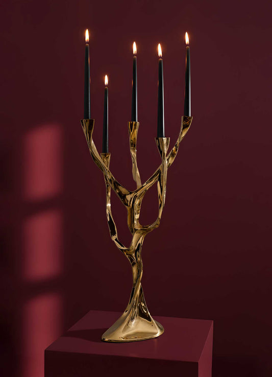 The Under The Banyan Gold Candelabra by VIYA, featuring five lit black candles and an organic banyan-inspired design, stands on a dark red pedestal against a deep red background, with shadows and reflections enhancing its sculptural beauty.