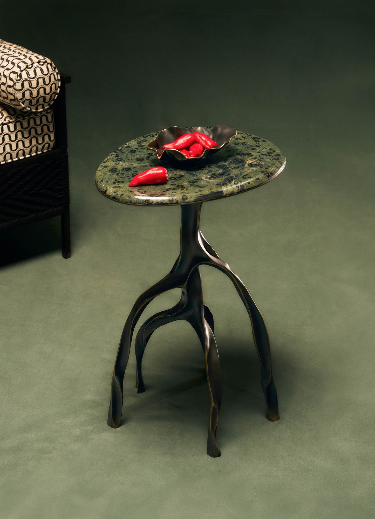 The VIYA Under The Banyan Side Table Large features a green marbled top and sculptural, banyan tree-inspired legs. On it sits a metal bowl with red chili peppers. A patterned armchair appears partially on the left.